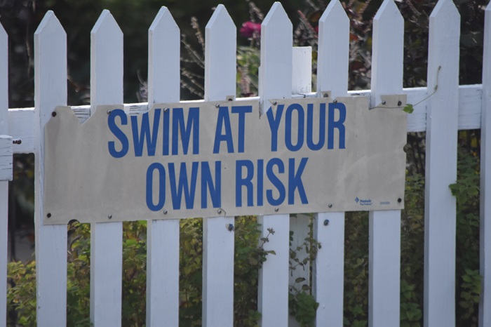 “Swim at Your Own Risk” warning sign attached to a white fence near a swimming pool area.
