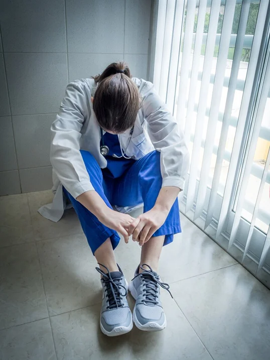 A doctor is sitting on the floor by a window with her head down. She looks tired.