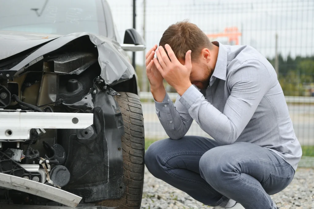 A man is crouched near his car, head in his hands. He is distressed after a car accident. 