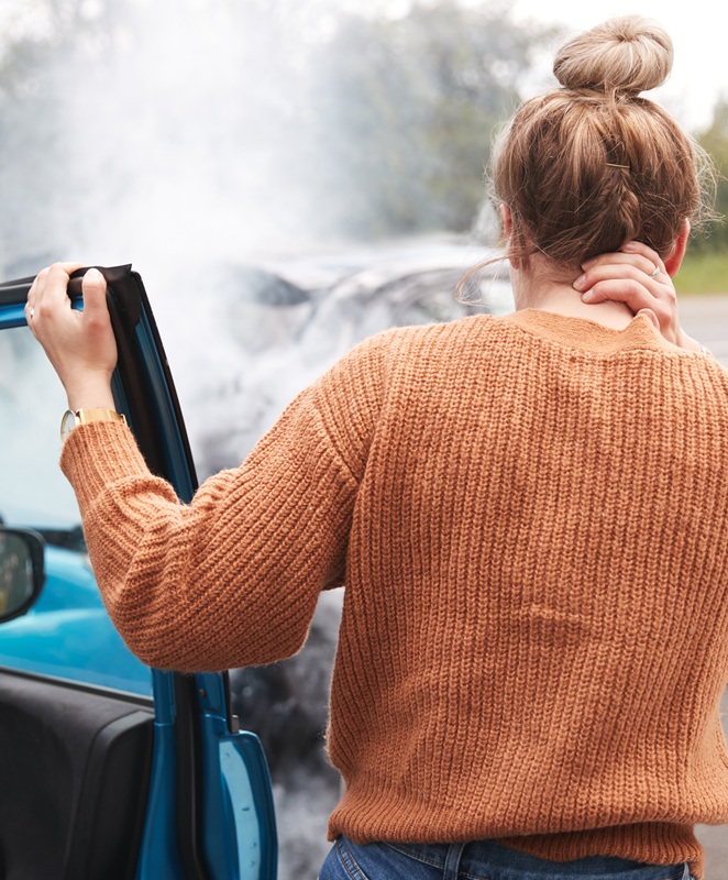 A woman is holding her neck, standing next to her car and looking at the aftermath of a bad accident.