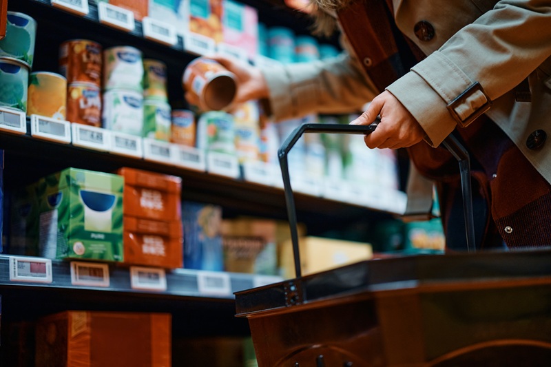 Closeup image of a woman grabbing a canned item off a grocery shelf.