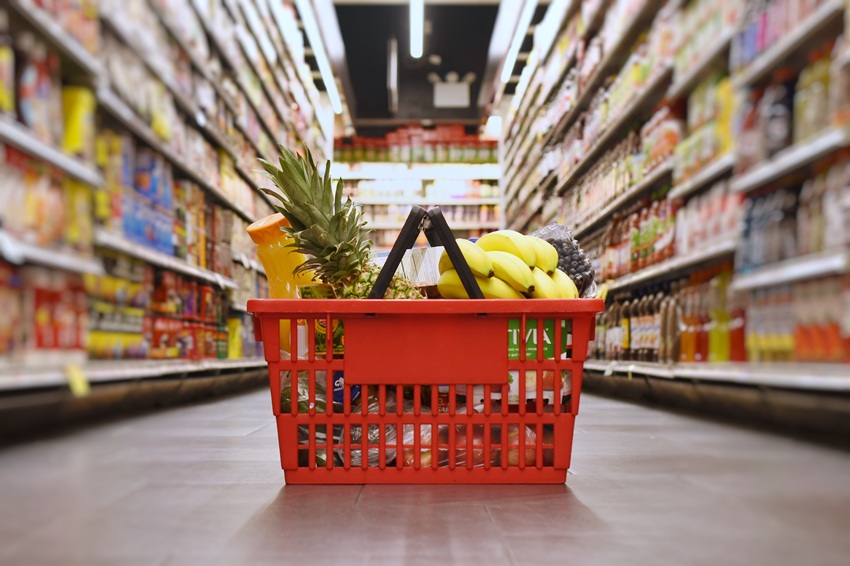 A red shopping handbasket filled with items. It is on the ground in an aisle, surrounded by groceries.